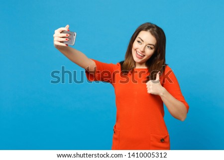 Beautiful brunette young woman wearing red orange dress doing selfie shot on mobile phone isolated over trendy blue background, studio portrait. People lifestyle fashion concept. Mock up copy space