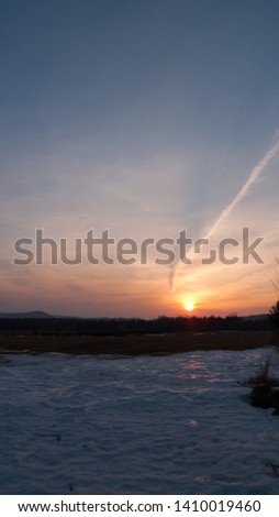 Picture taken at dusk in late winter with snow in the field, a forest in the back, a setting sun with high thinning clouds.  A vapor trail from an airplane creates a shadow on the clouds above.