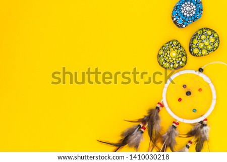 Dreamcatcher with brown bird feathers, and with threads and beads of rope hanging. Dreamcatcher handmade. It lies on a yellow background among the painted colored stones. There is a place for text.
