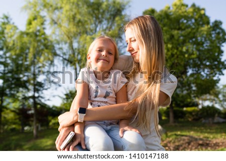 Beautiful young mother and daughter with blonde hair embracing outdoor. Stylish girls making walking in the park. Family concept
