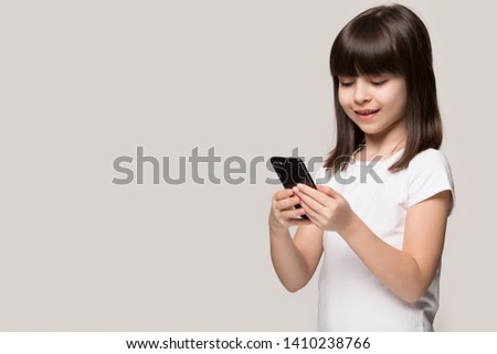 Little girl hold phone look at smartphone screen use mobile application chatting with friend, play game isolated on beige grey background copy space aside, parental control kid and modern tech concept