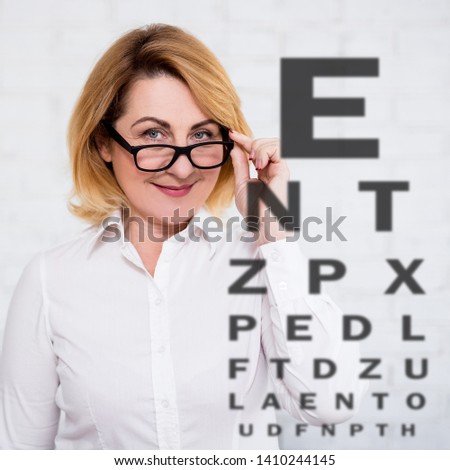 mature woman in eyeglasses and eyevision test chart over white wall background