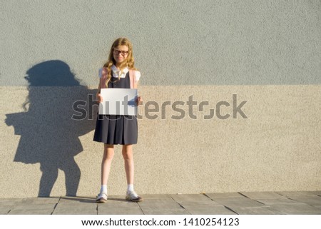 Little girl in school uniform with backpack showing a blank white sheet poster, outdoor wall background gray wall