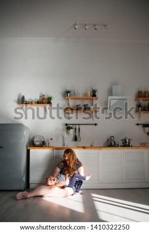 Mom and daughter are or sit on the floor of the bright kitchen. The girl is played with her mother.