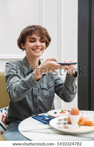 Attractive young girl having breakfast at the cafe indoors, taking a photo, blogging