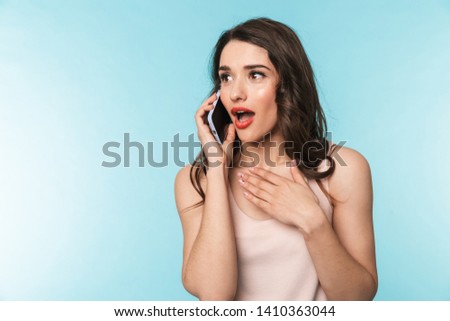 Portrait of a beautiful young brunette woman standing isolated over blue background, talking in mobile phone