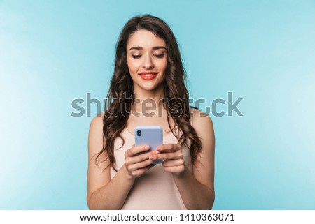 Portrait of a beautiful young brunette woman standing isolated over blue background, using mobile phone