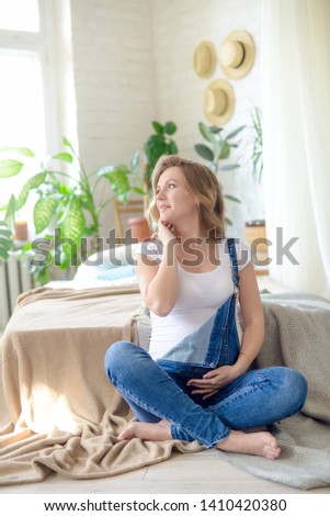 Beautiful pregnant woman with blond hair in a white T-shirt and blue jeans is sitting on a bed . Concept of happy motherhood, healthy lifestyle