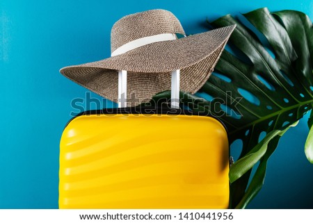 Bright and stylish cabin size suitcase with straw hat and monstera leaf against dark blue background. Easy travel with little baggage concept. Copy space.