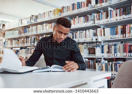 In stylish black shirt. African american man sitting in the library and searching for some information in the books.