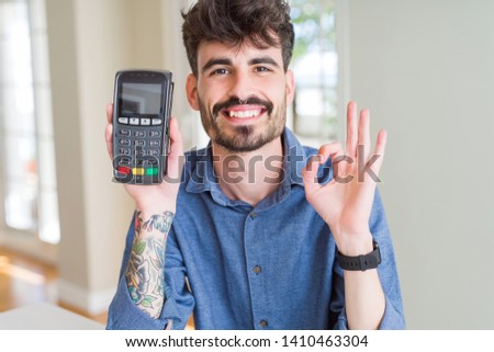 Young man holding dataphone point of sale as payment doing ok sign with fingers, excellent symbol