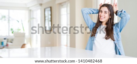 Wide angle picture of beautiful young woman sitting on white table at home Posing funny and crazy with fingers on head as bunny ears, smiling cheerful