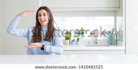 Wide angle picture of beautiful young woman sitting on white table at home gesturing with hands showing big and large size sign, measure symbol. Smiling looking at the camera. Measuring concept.