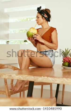 Healthy drink. Woman drinking coconut water in kitchen. Smiling young female in summer clothes enjoying green coconut milk at home
