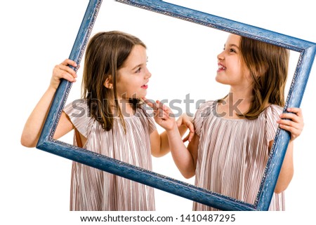 Identical twin girls are making happy expressions with picture frame. Children, sisters, girls posing in studio with picture frame, making different facial expressions. Family portrait, frontal view.