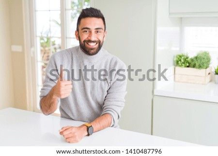 Handsome hispanic man wearing casual sweater at home doing happy thumbs up gesture with hand. Approving expression looking at the camera with showing success.