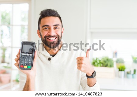 Handsome hispanic man holding point of sale terminal dataphone happy with big smile doing ok sign, thumb up with fingers, excellent sign