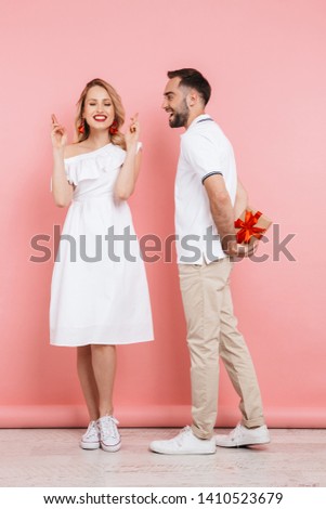 Full length of an attractive cheerful young couple standing together isolated over pink background, giving present, celebrating