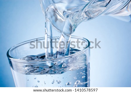 Pouring water from jug into glass on blue background Royalty-Free Stock Photo #1410578597
