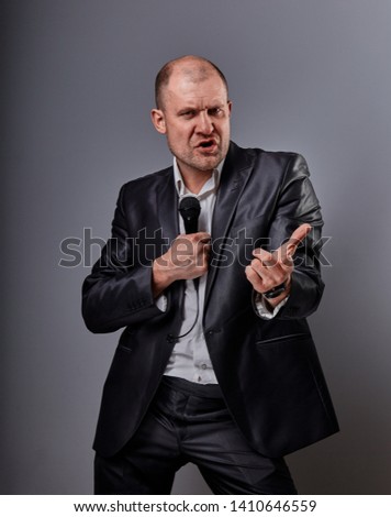 Fun emotional moving happy performer man presenting the show holding microphone in hand and showing the fist success sign on grey color background. Closeup portrait
