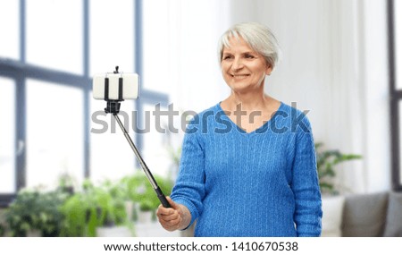 technology and old people concept - smiling senior woman taking picture by smartphone on selfie stick over home living room background