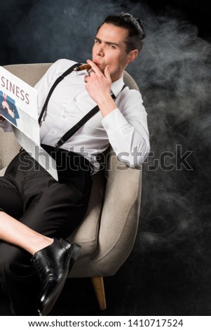 overhead view of handsome man sitting in armchair holding business newspaper and smoking cigar on black with smoke 