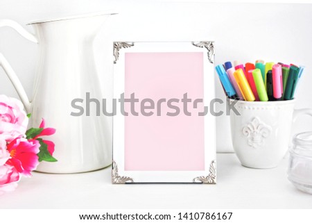 Empty pink frame with place for text on the pink wall and table. Flowers, candle, markers, vintage cup and pitcher. Girly artist style room interior. Template mock up for paintings or photographs.