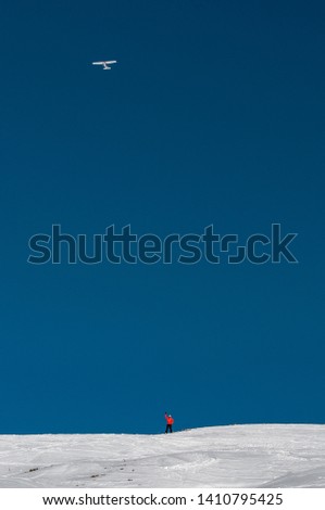 A snowboarder standing on the slope and an airplane in the blue sky flying above him