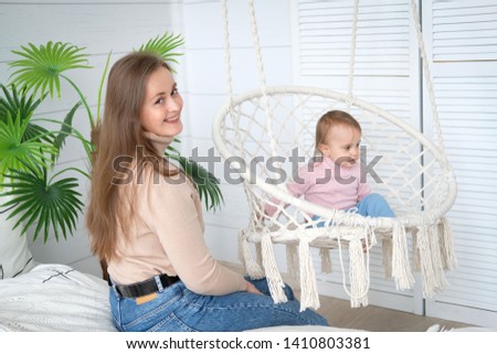 A little girl sitting in a hanging chair in the bedroom. The baby's mother sits beside him on the bed. Bright room in Scandinavian style. Macrame wicker chair, knitted blanket, straw pillow. Cozy home