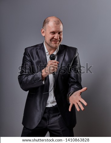 Fun emotional moving happy performer man presenting the show holding microphone in hand and showing the finger the choosing sign on grey color background. Closeup portrait
