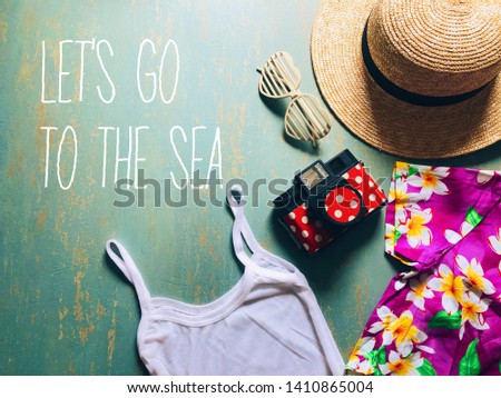 Summer theme straw boater hat with black ribbon, plastic heart shaped glasses, toy camera, white camisole, colorful hawaiian shirt on vintage green background with typography LET’S GO TO THE SEA.