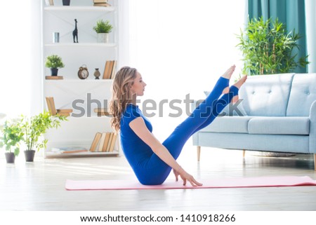 Young athletic girl fitness trainer in blue sports overalls shows morning gymnastic exercises in the interior of room. Stretching and swinging legs