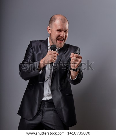 Smiling emotional moving happy performer man presenting the show holding microphone in hand and showing the success sign the fist on grey color background. Closeup portrait