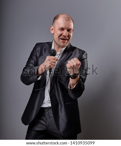 Smiling emotional moving happy performer man presenting the show holding microphone in hand and showing the success sign the fist on grey color background. Closeup portrait