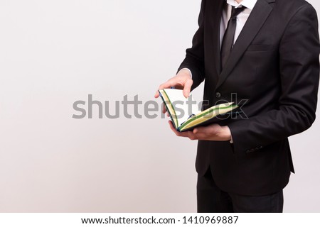 
A young man in a black jacket, white shirt and black tie is looking for something in a notebook