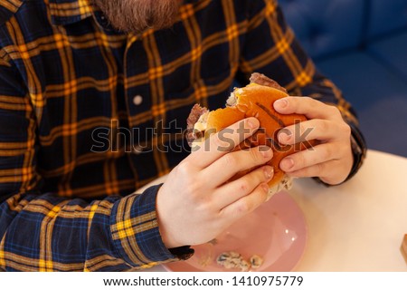Man in a tartan shirt is eating a burger with golden bun and grilled beef patty