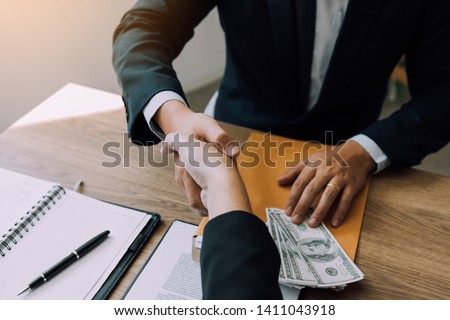 Two corporate businessmen shaking hands while one man places money on document in office room with corruption concept. Royalty-Free Stock Photo #1411043918