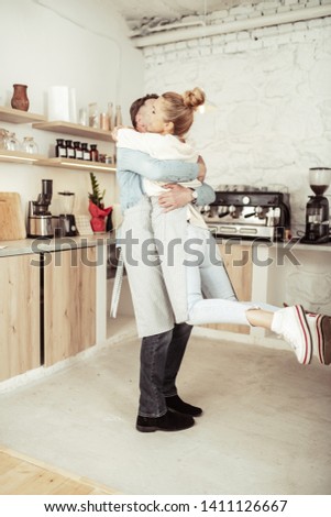 Love each other. Happy woman jumping in arms of her handsome husband in their kitchen.