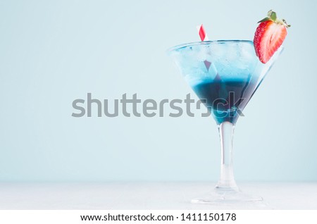 Sweet summer cold alcohol blue drink with ice cubes, red straw, strawberry in  wineglass on soft light pastel blue background.