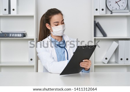 Woman doctor sits at the table in a medical mask and a stethoscope around his neck                  