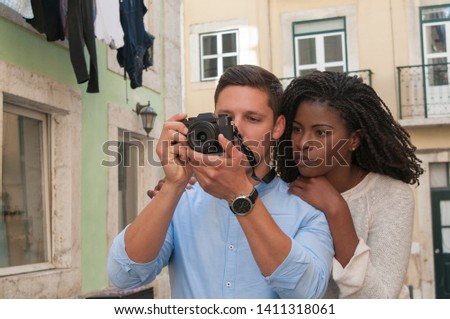 Serious interracial couple taking photos on camera in city. Woman standing and leaning on man shoulder in street with building in background. Romance and tourism concept. Front view.