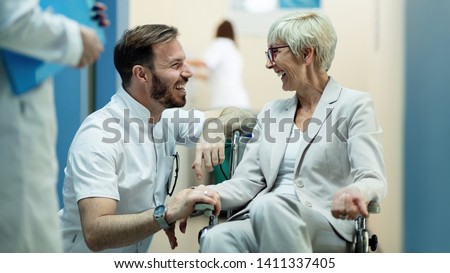 Happy male doctor communicating with mature patient who is in wheelchair in a hallway at clinic.   Royalty-Free Stock Photo #1411337405