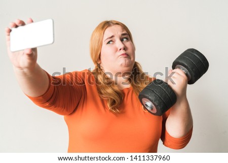 Chubby woman sport at home standing isolated on white wall holding smartphone taking selfie pictures with dumbbell grimace upset