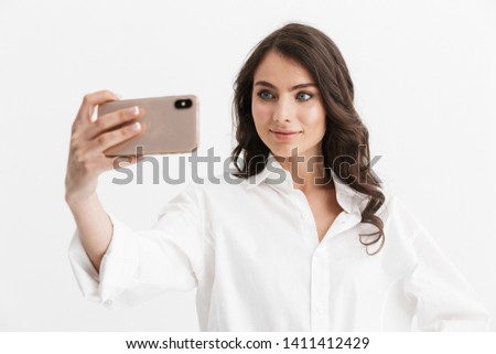 Beautiful excited young woman with long curly brunette hair wearing white shirt standing isolated over white background, taking a selfie