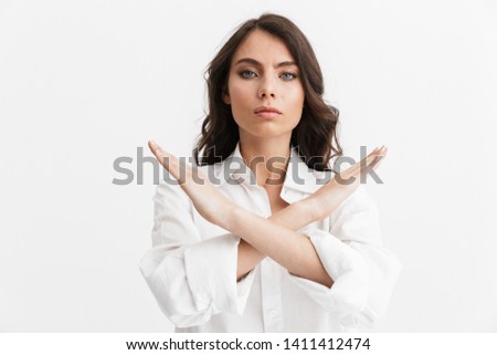 Beautiful angry young woman with long curly brunette hair wearing white shirt standing isolated over white background, showing stop gesture