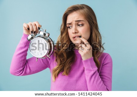 Beautiful shocked confused young girl wearing casual clothes standing isolated over blue background, showing alarm clock