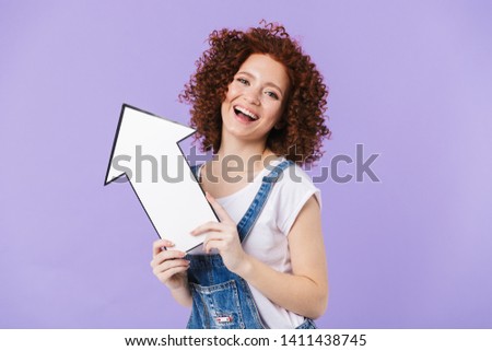 Image of a beautiful curly happy redhead girl posing isolated over purple background holding arrow.