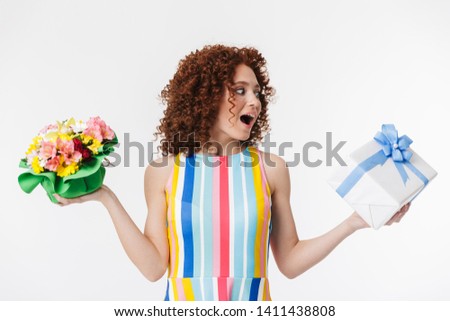 Portrait of cheery redhead curly woman 20s wearing colorful dress holding flowers and birthday present box isolated over white background