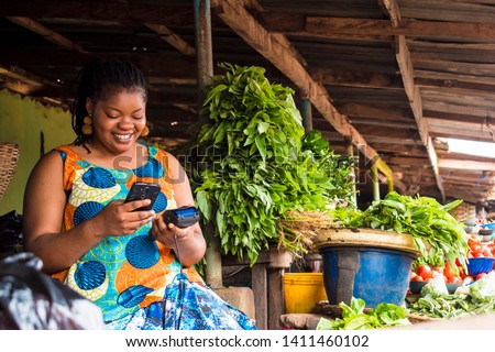 african woman using her phone and also holding a pos system in a local market Royalty-Free Stock Photo #1411460102