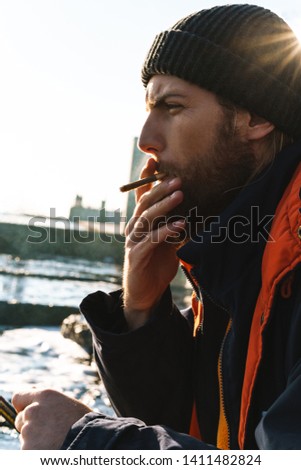 Picture of a handsome young man fisherman wearing coat and hat at the seashore smoking.